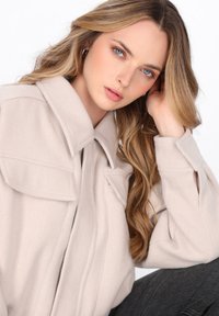 Lichtbeige oversized jas met kraag, twee borstzakken en een zachte textuur; model met lang, golvend haar tegen een effen achtergrond.