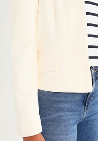 Blazer crema chiaro con un design strutturato, texture liscia e vestibilità su misura, abbinato a una maglietta a righe blu e bianche e jeans di denim.