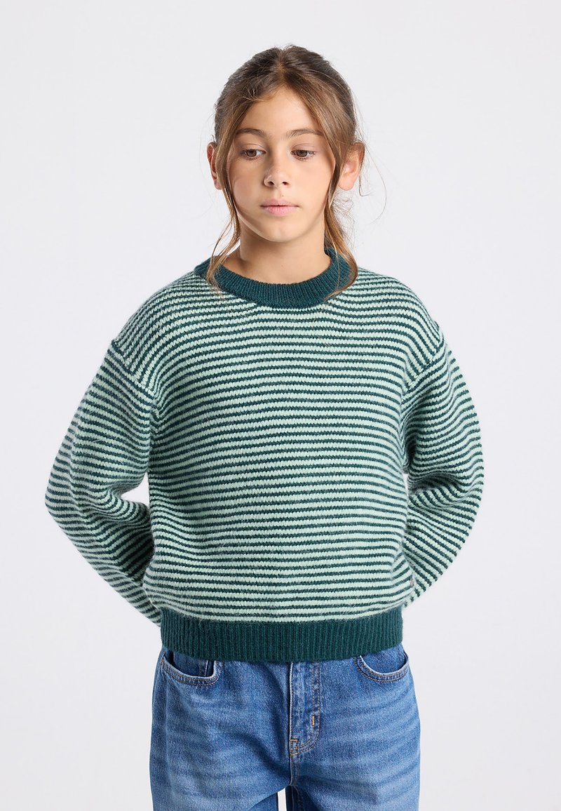 Maglione a righe con filato verde e bianco, caratterizzato da un collo rotondo, vestibilità ampia e bordi e orlo a costine. Abbinato a jeans in denim.
