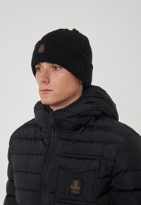Berretto lavorato a maglia nero con texture a coste e una patch con logo. Indossato con un giubbotto imbottito nero dotato di zip e tasca frontale.