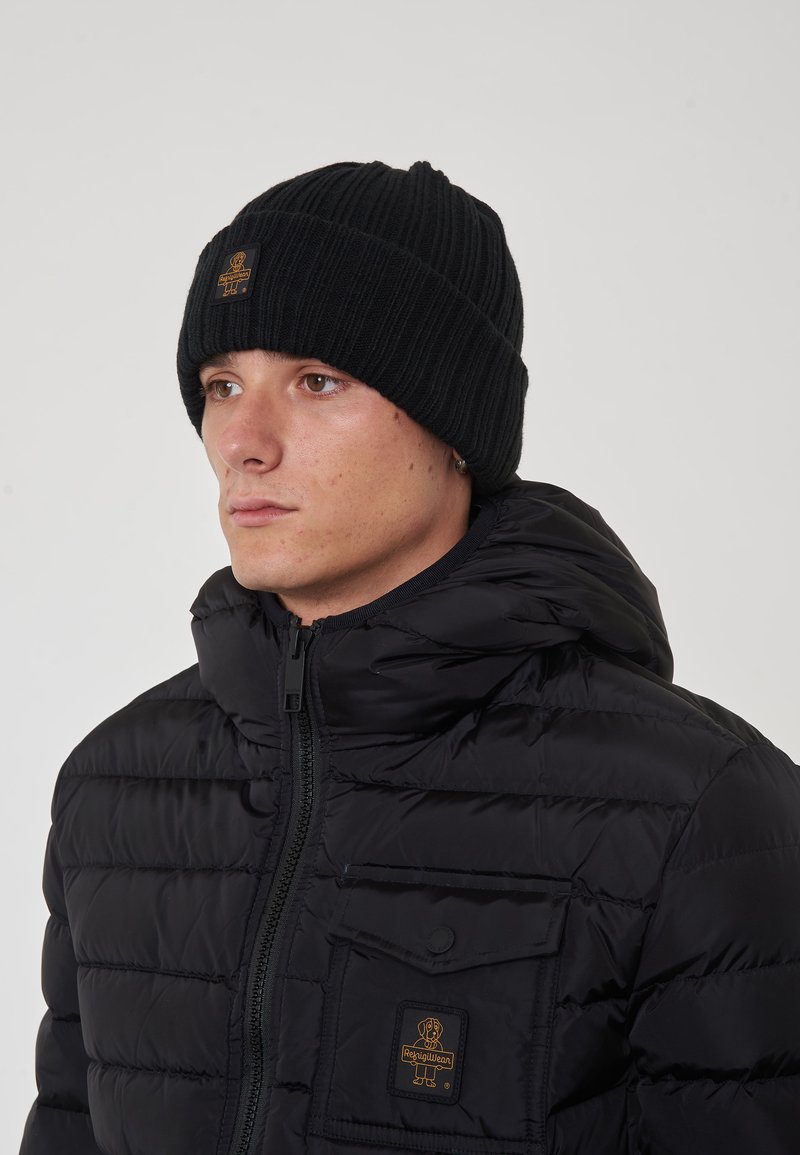 Berretto lavorato a maglia nero con texture a coste e una patch con logo. Indossato con un giubbotto imbottito nero dotato di zip e tasca frontale.