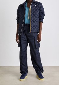 Giubbotto imbottito blu navy con logo bianco, abbinato a una maglietta teal e pantaloni cargo larghi. Le sneakers blu hanno una suola gialla. Texture liscia in tutto il capo.