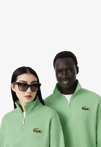Sudaderas verdes con cremallera y logo de cocodrilo bordado, que presentan cuellos altos y un ajuste relajado, llevadas por dos modelos.