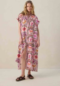 Robe rose à motifs tropicaux et texte, arrivant au genou avec des manches courtes et une fente latérale, assortie à des sandales marron foncé.