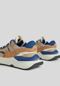 Pár béžových, modrých a bílých tenisek s detaily z prodyšné a kožené látky, tlustými bílými podrážkami a černým vzorovaným profilem na světle šedém pozadí.