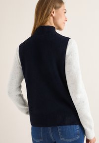 Marineblaue gestrickte ärmellose Weste mit strukturiertem Stoff, ausgestattet mit langärmeligen, hellgrau gefleckten Ärmeln und einem hohen gerippten Kragen.
