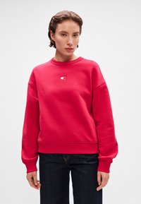 Sweatshirt rouge surdimensionné avec un col rond, des poignets côtelés et un petit logo brodé au centre. Tissu lisse avec une coupe décontractée.