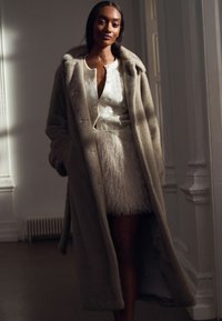 Lange, lichtbruine, zachte jas met een bontachtige textuur en een oversized kraag, gedragen over een wit hemd met knopen en een crèmekleurige, gestructureerde rok.