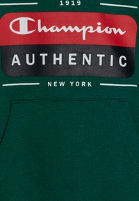 Sudadera de algodón verde con un gran logo de Champion en rojo y negro, texto blanco que dice "AUTÉNTICO" y "NUEVA YORK", y un bolsillo frontal.