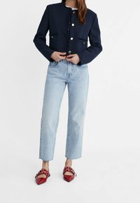 Veste marine à col avec détails en franges, boutons dorés, associée à un jean droit bleu clair et des chaussures rouges à bout pointu avec des accents de lanières.