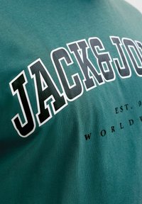 Zeleni materijal s istaknutim crnim tekstom obrubljenim bijelom bojom "JACK & JONES" i manjim crnim tekstom djelomično vidljivim ispod.