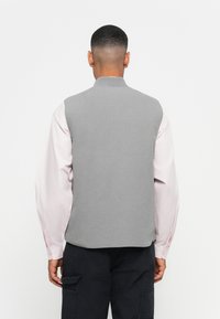 Άνδρας με κοντά μαλλιά που φοράει γκρι без рукава πάνω από ένα ανοιχτό ροζ μακρυμάνικο πουκάμισο και μαύρα cargo παντελόνια, γυρισμένος μακριά από την κάμερα.