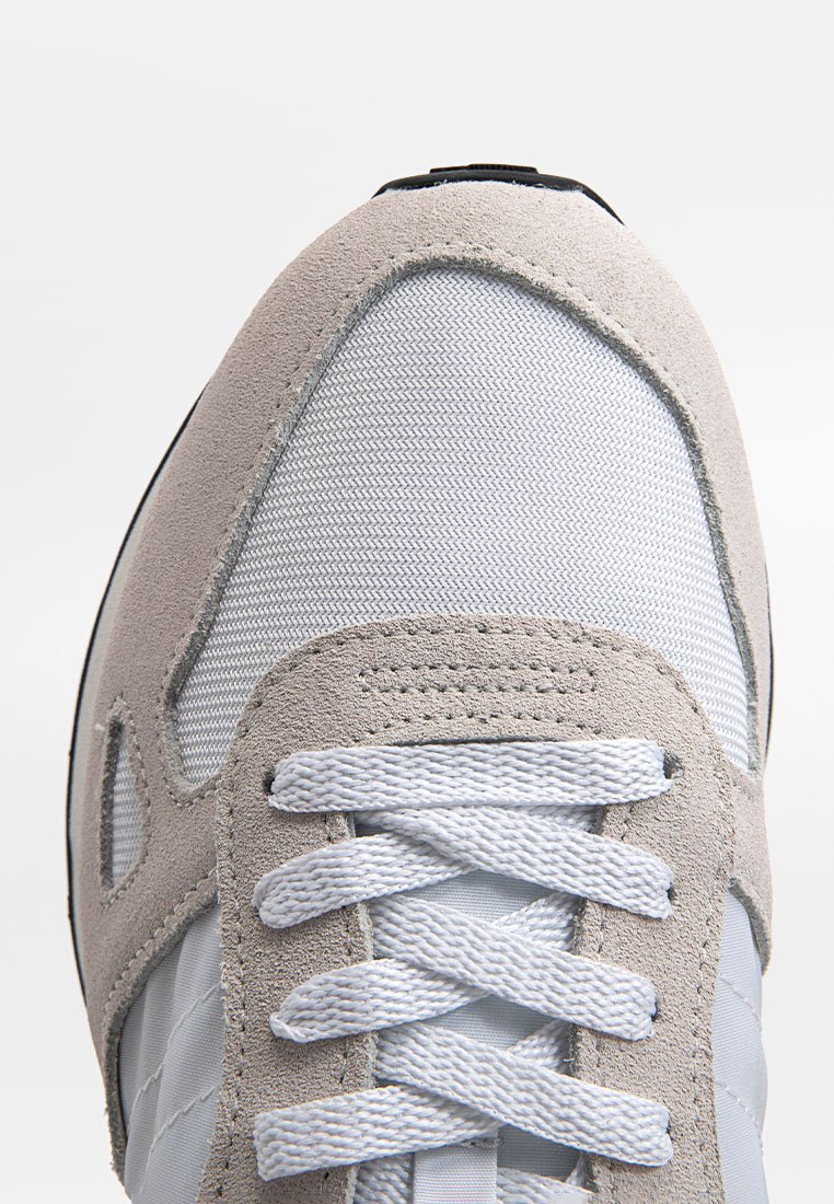 Zapato deportivo de color gris claro que presenta materiales de ante y malla, puntera redonda, cordones blancos y costuras detalladas en la parte superior.