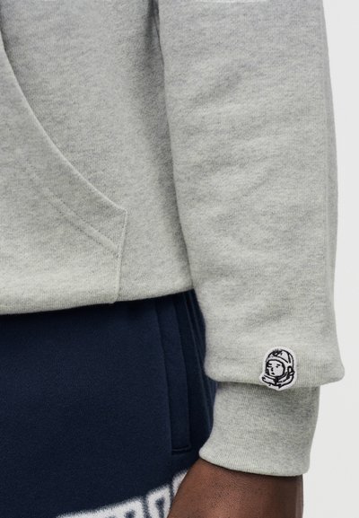 Γκρι φούτερ με απαλή υφή, με μικρό κεντημένο λογότυπο στο μανίκι και μπροστινή τσέπη. Συνδυάζεται με σκούρο μπλε παντελόνι φόρμας.