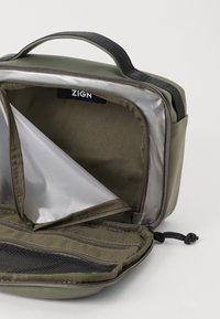 Piccola borsa da viaggio verde oliva con zip e manico, aperta per mostrare l'interno con rivestimento simile alla seta, tasca in rete e etichetta del marchio "ZIGN."