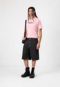 Rožinė medvilninė marškinėliai su juodu "Pepe Jeans" logotipu, derinami su tamsiais džinsiniais šortais ir juodais bateliais su raišteliais. Paprastas dizainas, laisvas siluetas.