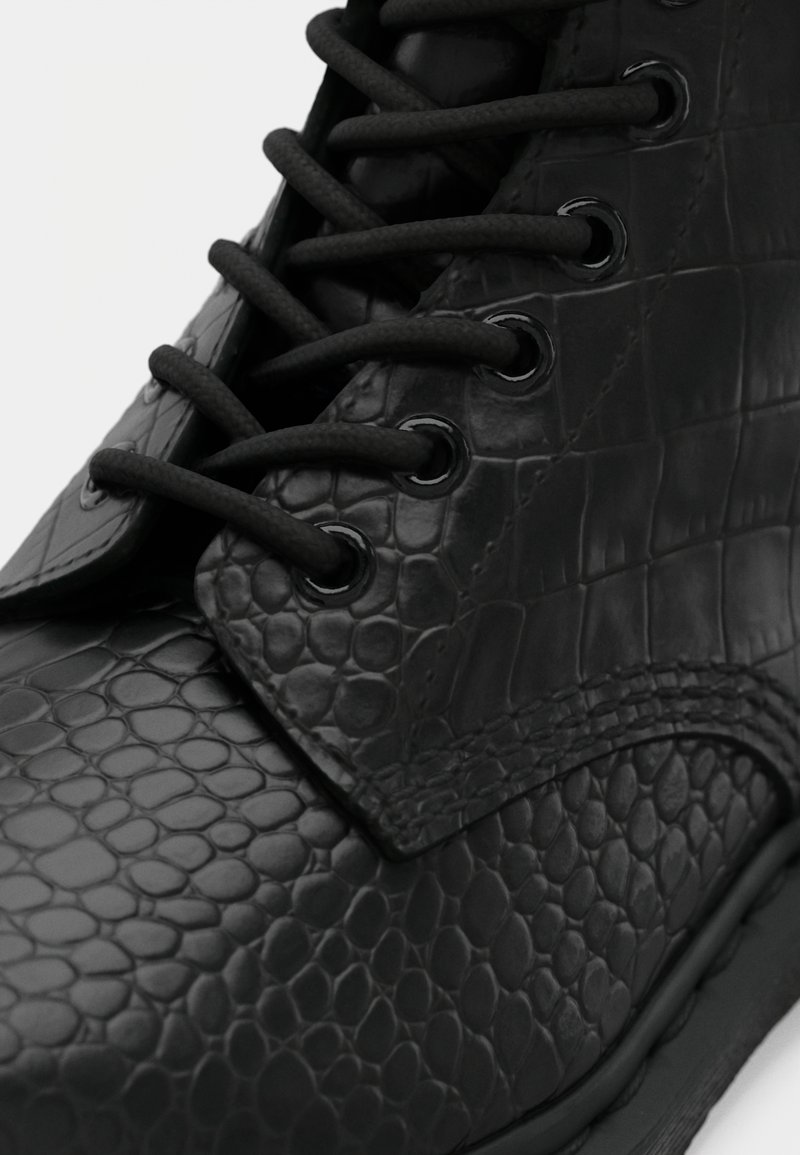 Bota negra con cordones, confeccionada en cuero texturizado con un patrón similar al de un reptil y ojales de metal negro, mostrada en un primer plano detallado.