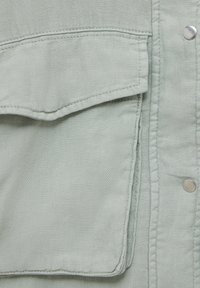 Tissu vert clair en gros plan présentant une poche, des coutures visibles et des boutons-pression en métal. La texture semble douce avec un léger éclat.