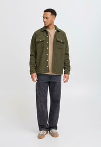 Dunkelgrüne Fleece-Shirtjacke mit zwei Brusttaschen, Knopfverschluss und einem leicht lockeren Schnitt über einem beigen Oberteil und dunklen Jeans.