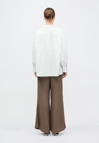 Barna haját kontyba fogó nő laza, fehér, hosszú ujjú ingben és bő szárú barna nadrágban áll egy egyszerű, fehér háttér előtt.