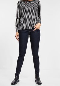 Černobílý top s dlouhým rukávem a vzorem na zig-zag s kulatým výstřihem, v kombinaci s tmavými slim-fit džíny a kotníkovými botami.