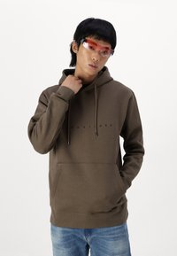 Brun hætte sweatshirt med en frontlomme og sort tekst. Modellen bærer orange-farvede solbriller og blå jeans mod en hvid baggrund.