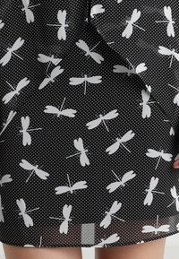 Svart tyg med vita prickar och trollslände-mönster. Lätt textur med en halvgenomskinlig kvalitet. Fyrkantig lappdetalj nära fållen.