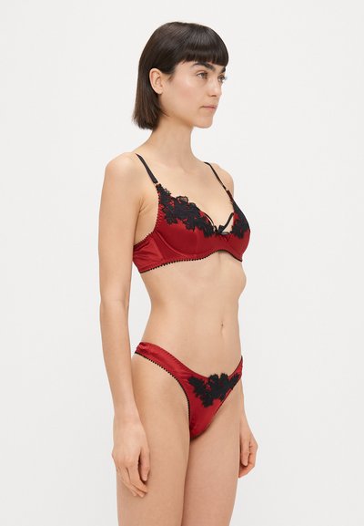 Červený saténový spodný prádlový set s čiernymi čipkovými akcentmi, bralette s nastaviteľnými ramienkami a zodpovedajúci string s zložitými čipkovými detailmi.