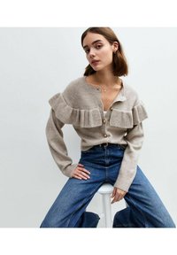 Beige stickad kavaj med volangkrage, prydd med guldknappar, i kombination med blå vida byxor. Mjuk textur, avslappnad passform och lagerdesign.