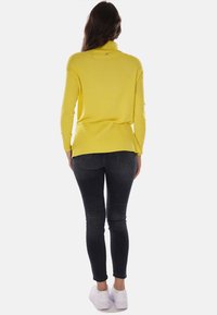 Maglione a maniche lunghe con colletto alto a coste di un brillante colore giallo, abbinato a jeans skinny neri e sneakers bianche, visto da dietro.