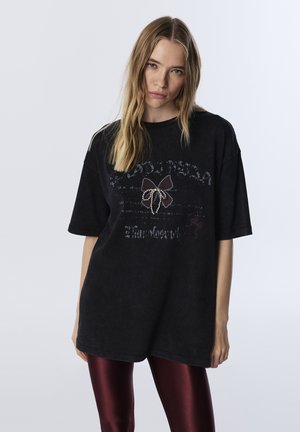 Schwarzes, oversized T-Shirt mit strukturiertem Design und dekorativem Bogen in der Mitte, kombiniert mit glänzenden burgunderfarbenen Leggings.
