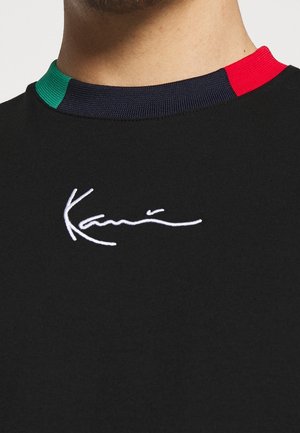 Czarna koszulka z okrągłym dekoltem, z białym logo pisanym kursywą na środku klatki piersiowej oraz kołnierzem w blokach kolorów zielonego, granatowego i czerwonego.