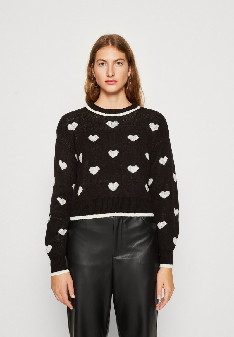 Črn skrajšan pulover z belim vzorcem srčkov, rebrastimi manšetami in ovratnikom. Model ga nosi z črnimi usnjenimi hlačami na nevtralnem ozadju.