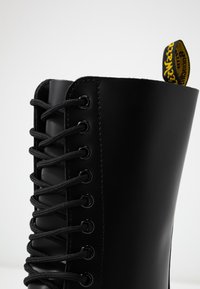 Close-up of a black lace-up leather boot with a yellow and black fabric pull tab at the top.