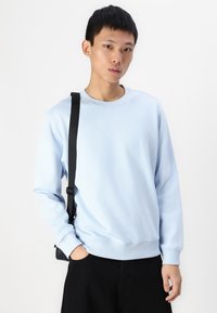 Light blue sweatshirt in soft cotton blend, featuring a round neckline and long sleeves. Paired with black pants and a shoulder bag.