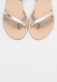 Sandaalit, joissa on pehmeä beige pohja ja vihreät ja hopeiset metalliset hihnat, jotka risteävät päällä. Sujuva pinta ja minimalistinen muotoilu.