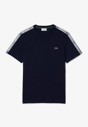 Granatowy t-shirt z krótkim rękawem z białą taśmą z logo Lacoste na ramionach oraz małym zielonym logo krokodyla na lewej piersi.