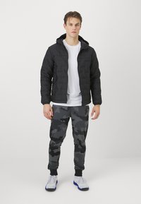 Giacca nera imbottita con cappuccio, maglietta bianca sotto e pantaloni sportivi grigi usati per il camouflage. Presenta polsini elastici e sneakers bianche.