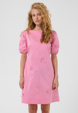Jeune femme aux cheveux blonds bouclés portant une robe rose mi-longue avec des manches bouffantes et de petits détails de nœuds.