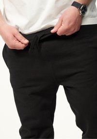 Black jogger pants with a drawstring waistband, featuring a smooth texture and fitted design. A person's hands adjusting the waistband are visible.