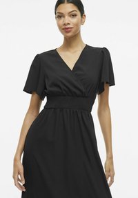 Must musträtik, millel on V-kaelus ja lühikesed varrukad, smockitud vöökoht ja voolav seelik lõdva istuvuse jaoks. Sile kangamaterjal.