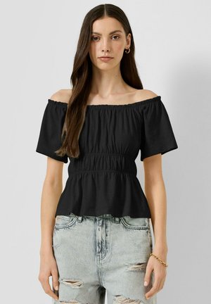 Jeune femme aux longs cheveux foncés portant un top noir décolleté aux épaules et un jean bleu clair déchiré, debout devant un fond uni.
