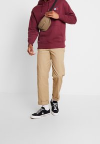 Bordowy hoodie, beżowe chinosy i czarne trampki z białymi akcentami. Brązowa torba na ramię. Luźny strój uliczny.