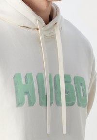 Sweat à capuche crème en tissu doux avec une grande inscription verte "HUGO" et des cordons à embouts blancs. Coupe classique avec capuche.