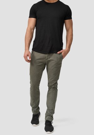 Homme portant une chemise noire à manches courtes, un pantalon ajusté vert olive les mains dans les poches, et des baskets noires marchant en avant.