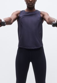 Άτομο με σκούρο αθλητικό τοπ και κολάν κρατά βαράκια 1 κιλού, τεντωμένα μπροστά, εκτελώντας άσκηση ενδυνάμωσης σε απλό φόντο.