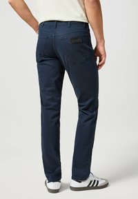 Chinos azul marino con un corte ajustado, que cuentan con un parche de cuero en el bolsillo trasero y detalles de costura sutiles. Tejido suave y duradero.