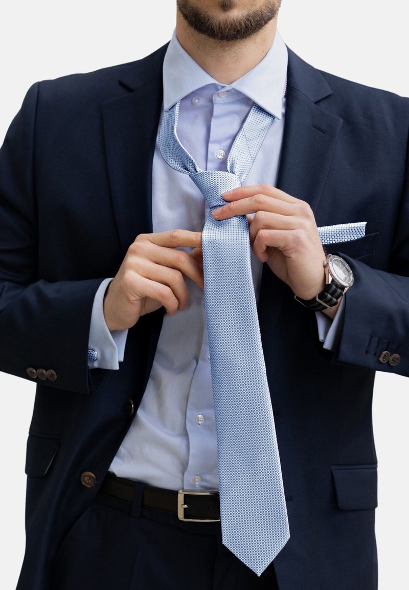 Cravatta blu chiaro texturizzata con una forma stretta, indossata in modo rilassato su una camicia blu chiaro, abbinata a un abito scuro e cintura nera.