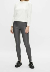 Weißes Longsleeve-Oberteil mit Puffärmeln, kombiniert mit grauen Fake-Leder-Leggings. Das Model trägt schwarze Schnürschuhe. Neutraler Hintergrund.