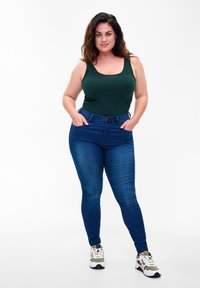 Tume roheline tank top koos tumehallide kitsaste teksadega. Teksadel on nup closure ja sujuv denim tekstuur. Modell kannab tossusid.